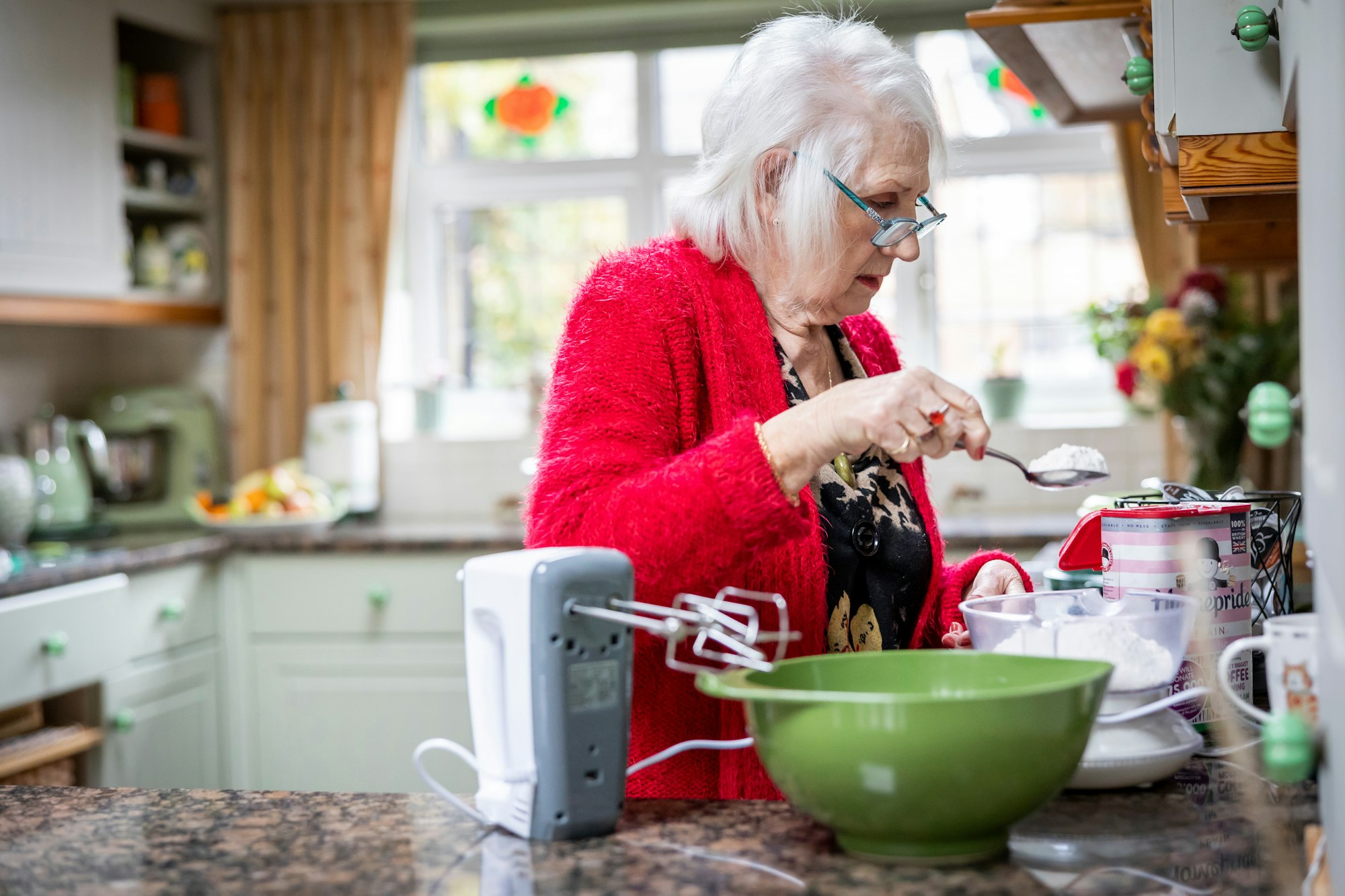 An older woman in a red cardigan cooking in a bright home kitchen.